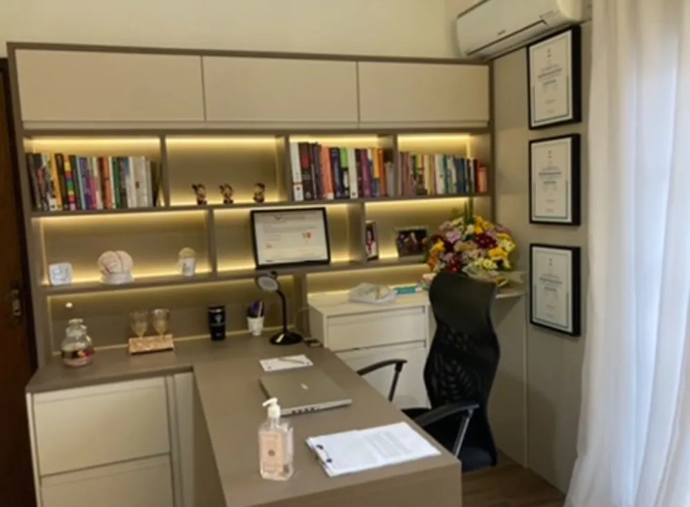 Consultório com mesa em L, estante iluminada com livros e flores
